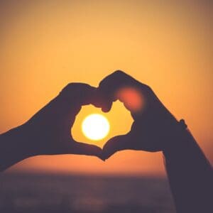 A person making a heart with their hands at sunset.